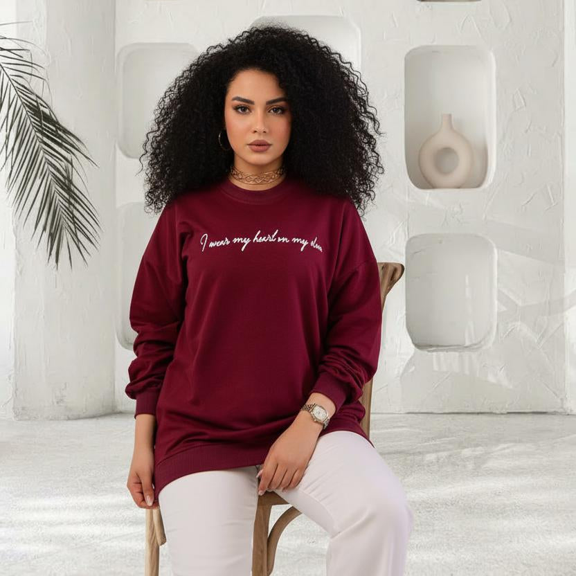 Woman wearing a burgundy sweatshirt with text, sitting in a modern indoor setting with white walls and decorative elements.